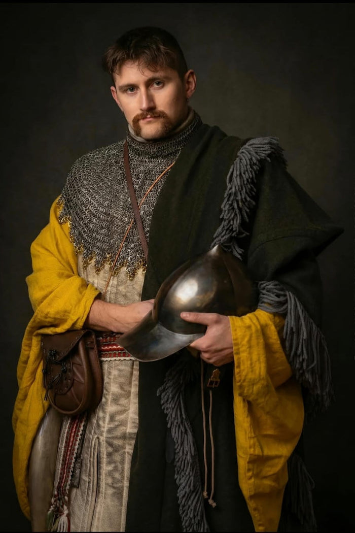 Peter Tobin afgebeeld als een 15e-eeuwse Ierse gallowglass-krijger, gekleed in een maliënkraag, gele léine en wollen brat, terwijl hij een middeleeuwse helm vasthoudt tijdens een historische reenactment fotoshoot.