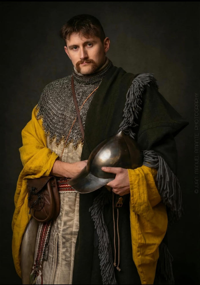 Peter Tobin afgebeeld als een 15e-eeuwse Ierse gallowglass-krijger, gekleed in een maliënkraag, gele léine en wollen brat, terwijl hij een middeleeuwse helm vasthoudt tijdens een historische reenactment fotoshoot.